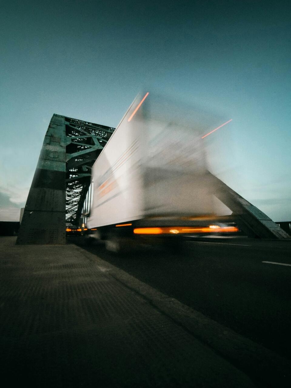 Motion-blurred dry van trailer crossing a steel truss bridge at dusk