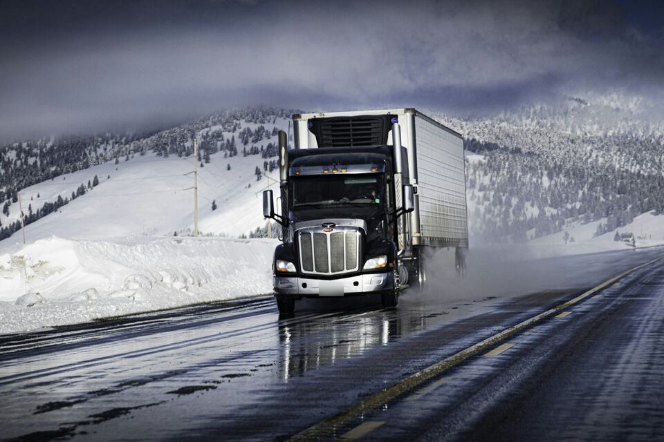 Long-nose tractor pulling a refrigerated trailer on a wet highway between snowy hills